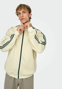 Jeune homme fermant sa veste de survêtement beige avec des bandes vert foncé sur les manches, portant un pantalon marron clair, sur fond blanc uni.