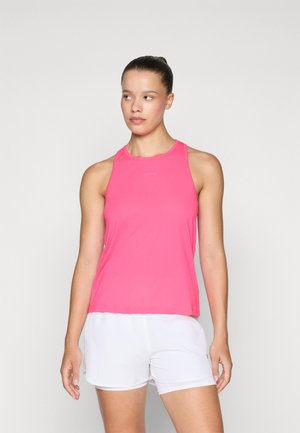 Kvinne med håret i en knute, iført en lys rosa ermeløs treningsgenser og hvite shorts, som står foran en ensfarget lys bakgrunn.