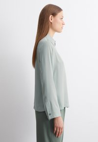 Light green blouse with a collared neckline, long sleeves, and button accents on the cuffs. Smooth, flowing fabric and relaxed fit.