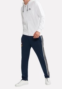 Vit zip-up hoodie med dragsko, marinblå joggers med vita ränder och logotypdetaljer, i kombination med vita sneakers.