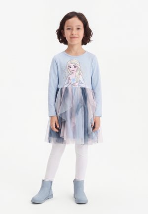 Enfant aux cheveux courts et foncés portant une robe bleue Frozen avec une jupe en tulle, des leggings blancs et des bottes bleues, debout devant un fond uni.