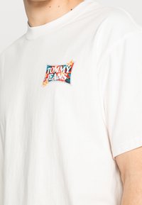 Camiseta de algodón blanca con un parche colorido del logo "Tommy Jeans" en la parte izquierda del pecho. Mangas cortas, cuello redondo, corte casual.