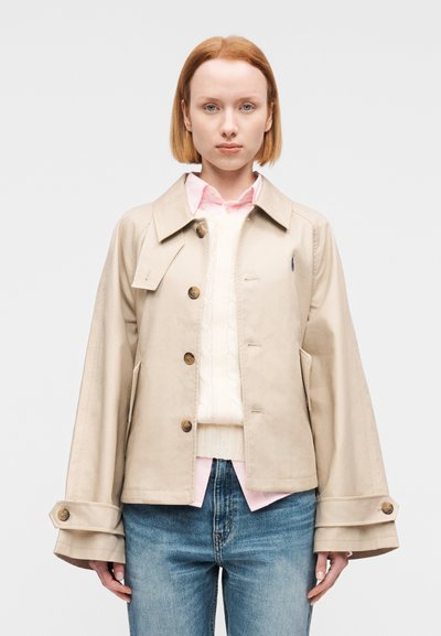 Μπεζ κοντό μπουφάν από βαμβάκι με φαρδιά γιακά, τέσσερα κουμπιά και δύο πλαϊνές τσέπες. Συνδυασμένο με λευκό πουλόβερ με πλέξη καλώδιο και ροζ πουκάμισο.