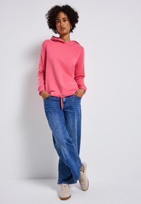 Felpa rosa con cappuccio e cordoncino davanti, maniche lunghe con strisce bianche, abbinata a jeans denim blu larghi e sneakers beige.
