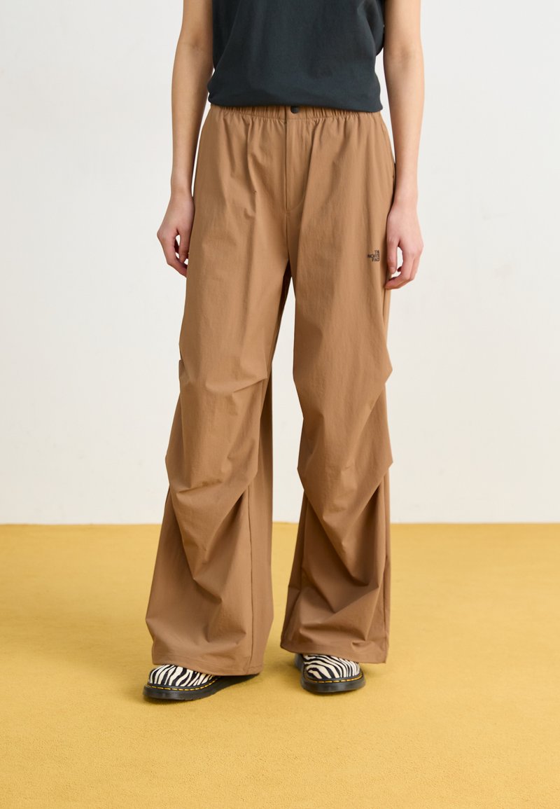Pantalons amples de couleur marron en tissu léger, dotés d'une taille élastique, avec des plis prononcés et un logo de marque près de la poche.