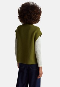 Osoba s kudrnatými vlasy, oblečená v olivově zeleném svetru bez rukávů přes bílý texturovaný top s dlouhým rukávem a tmavě modrých kalhotách, otočená zády.