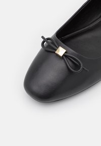 Zwarte leren platte schoen met een ronde teen, voorzien van een decoratieve strik en een goudkleurig vierkant accent op de bovenkant. Gladde textuur.