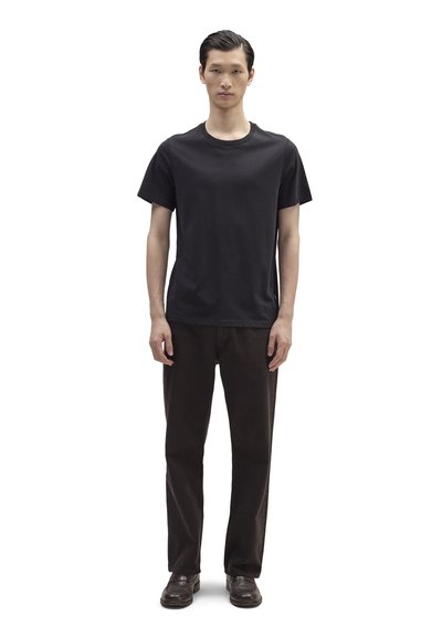 Νεαρός άνδρας που στέκεται και κοιτάζει προς τα εμπρός, φοράει μαύρο κοντομάνικο μπλουζάκι, σκουρόχρωμο παντελόνι και σκουρόχρωμα παπούτσια, με φόντο λευκό.