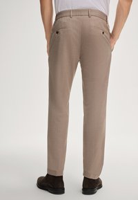 Beige op maat gemaakte broeken met een textuurpatroon, voorzien van achterzakken met knopen en een slim fit. Draagt een paar donkerbruine suède schoenen.