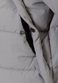 Tuvumā redzams pelēcīgs polsterēts apģērbs ar atvērtu pogu kabatu, kurā redzams melns audums kabatas iekšpusē.