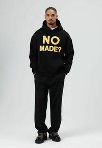 Zwarte hoodie met gouden tekst "NO MADE?" en bijpassende zwarte broek. Model draagt chunky zwarte schoenen met roze veters, staand tegen een lichte achtergrond.