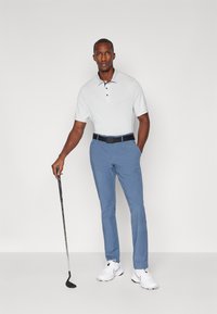 Golfbekleidung mit einem hellgrauen Polohemd mit dezentem Muster, blauen Hosen und weißen Schuhen, ergänzt durch einen schwarzen Gürtel und einen Golfschläger.