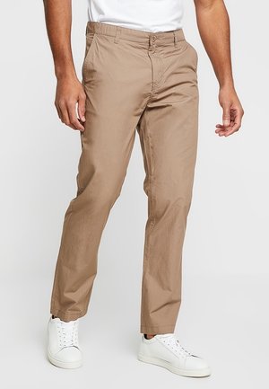 Mann trägt beige gerade geschnittene Hose und weiße Turnschuhe, steht vor einem einfarbigen hellen Hintergrund.