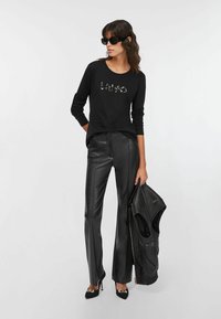 Top nero a maniche lunghe con logo "HUGO" in cristalli, abbinato a pantaloni neri in ecopelle a vita alta e décolleté nere con punta.