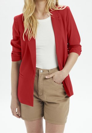 Röd, strukturerad kavaj med uppvikt ärm över en vit ribbad topp och beige shorts. Har en enkel knappstängning och sidofickor.