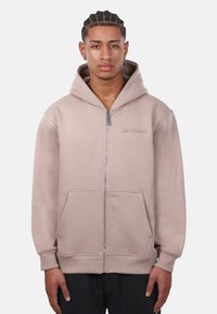 Haut de hoodie zippé beige clair avec capuche à cordon, poches avant et poignets côtelés. Logo brodé subtil sur la poitrine gauche.