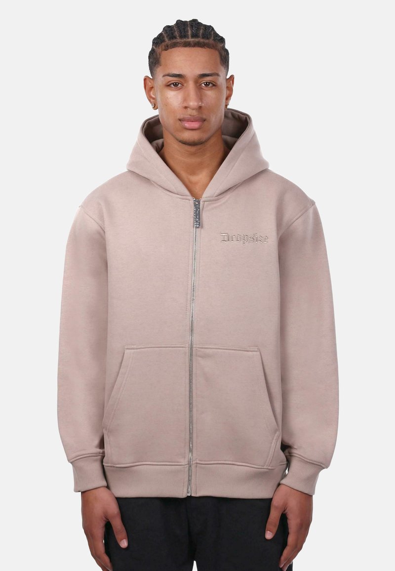 Haut de hoodie zippé beige clair avec capuche à cordon, poches avant et poignets côtelés. Logo brodé subtil sur la poitrine gauche.