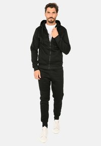 Felpa nera con zip e un cappuccio ampio, abbinata a joggers neri coordinati e sneaker bianche. Tessuto dalla superficie liscia.