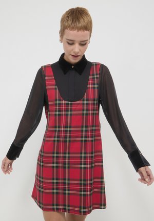 Abito scozzese rosso con scollo rotondo, realizzato in tessuto intrecciato. Presenta un design senza maniche, completato da una camicia nera trasparente a maniche lunghe.