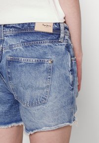 Vaaleansiniset farkkushortsit, joissa on repaleiset lahkeet, päällä henkilö, farkuissa on näkyvä takatasku ja beige Pepe Jeans -brändilogo.