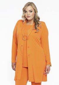 Orangenes dreiteiliges Outfit mit einem langen, offen getragenen Blazer, passendem Oberteil und enganliegenden Hosen. Glattes, leichtes Material ohne Muster.