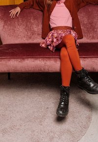 Dítě v růžovém tričku, vzorované sukni a oranžových legínách, sedí na růžové pohovce s černými šněrovacími botami a texturovaným béžovým kobercem.