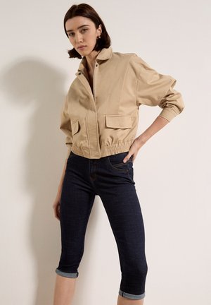 Mujer con chaqueta corta beige con bolsillos y jeans ajustados oscuros remangados, posando contra una pared blanca lisa.