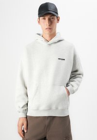 Młody mężczyzna ubrany w jasnoszary hoodie Abercrombie & Fitch, ciemne spodnie cargo i czapkę w kolorze czarnym, stojący na białym tle.