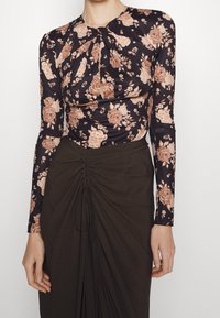 Maglietta nera a maniche lunghe con un motivo floreale in rosa e beige, caratterizzata da un design attorcigliato sul collo, abbinata a una gonna verde oliva arricciata.