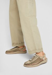 Beige footwear with a woven upper design, open heel, and rubber sole. Features include lacing and a flexible, textured surface.