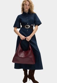 Bordeaux læder taske med et afslappet design, glat tekstur og bred åbning, holdt i hænderne, båret med en denim skjortekjole og bælte.