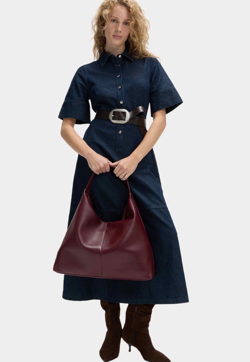 Bordeaux læder taske med et afslappet design, glat tekstur og bred åbning, holdt i hænderne, båret med en denim skjortekjole og bælte.