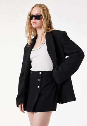 Blondynka w czarnych okularach przeciwsłonecznych, oversize'owej czarnej marynarce, białym topie na ramiączkach i czarnej spódnicy z wysokim stanem i guzikami, stojąca z ręką na biodrze.