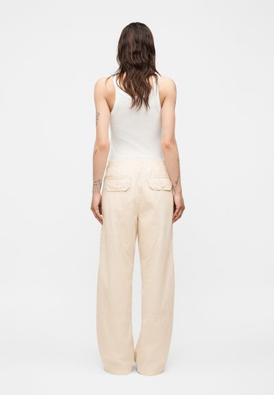 Γυναίκα με μακριά καστανά μαλλιά, φοράει λευκό αμάνικο τοπ και μπεζ φαρδιά παντελόνια, στέκεται με την πλάτη προς ένα απλό φόντο.