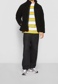 Person i svart fleecejacka över en gul-och-vit randig skjorta, lösa svarta byxor och vita sneakers, står mot en enkel bakgrund.