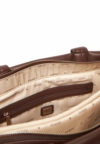 Intérieur d'un sac à main en cuir marron avec doublure en tissu présentant un logo "GUESS" en répétition et plusieurs compartiments, dont une poche zippée.