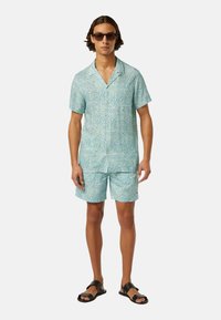 Light blue short-sleeve shirt and matching shorts with a geometric pattern. Model wears black sunglasses and sandals, standing on a plain background.