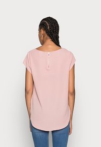 Kvinna med flätat hår iförd en ljusrosa kortärmad topp med dragkedja bak, kombinerat med blå jeans, stående mot en enfärgad bakgrund.