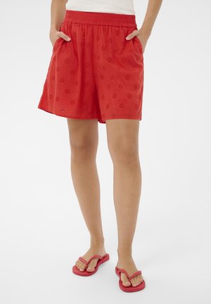 Person iført røde shorts med øjnelet broderi og elastisk talje, samt røde klipklappere, stående med hænderne i lommerne.