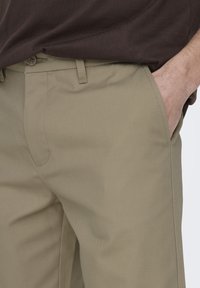 Beżowe spodnie wykonane z trwałej tkaniny, z płaskim przodem, bocznymi kieszeniami oraz dopasowanym krojem. Starannie wszyte, o gładkiej teksturze.