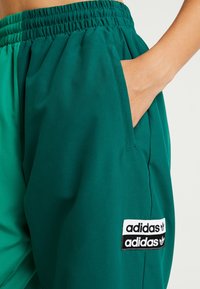 Gröna polyesterjoggers med elastisk midja och sidofickor, med en svart och vit Adidas-logotyp på nedre benet.