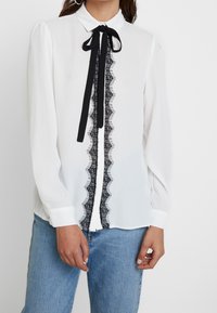 Camicia bianca con dettagli in pizzo nero lungo il centro e un fiocco al colletto. Maniche lunghe, tessuto morbido e polsini con bottoni.