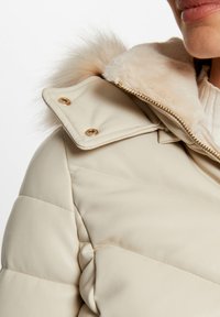Giubbotto puffer beige con collo in pelliccia sintetica, caratterizzato da cuciture diagonali, accessori in tonalità oro e una texture in pelle liscia.