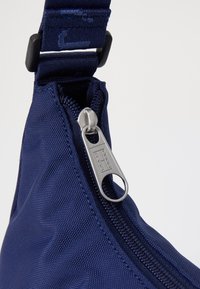 Sac zippé bleu marine avec une surface en nylon texturé. Présente un tirette en métal argenté et un régulateur de sangle en plastique. Design compact.
