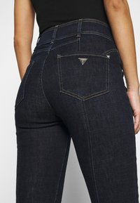 Jeans de denim azul oscuro con un diseño ajustado, cintura alta y costuras amarillas en contraste. Presentan un pequeño parche con el logo en el bolsillo trasero.