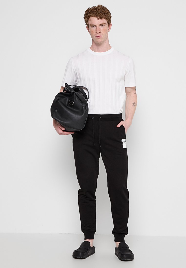 Schwarze Jogginghose mit Bündchen, ein weißes geripptes Rundhals-T-Shirt und eine schwarze Ledertasche mit glänzendem Hardware-Detail.