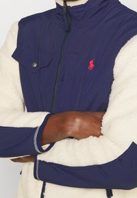 Tmavě modrá a krémová bunda s zapínáním na zip, kapsou na patentní knoflík a měkkou, nadýchanou texturou na těle s kontrastními rukávy.