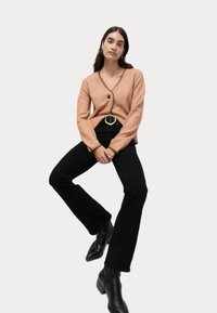Lys brun cardigan med V-hals, sorte knapdetaljer og kontrasterende kanter; parret med højtaljede sorte flared jeans og sorte støvler.