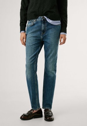 Persoon die blauwe rechte jeans, zwarte gestudloafers, een zwarte trui en een lichtblauw overhemd draagt met mouwen die zichtbaar zijn bij de polsen.
