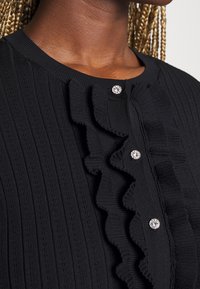 Pull noir côtelé avec des volants verticaux et trois boutons ronds scintillants, porté par une personne aux cheveux tressés et à la peau foncée.